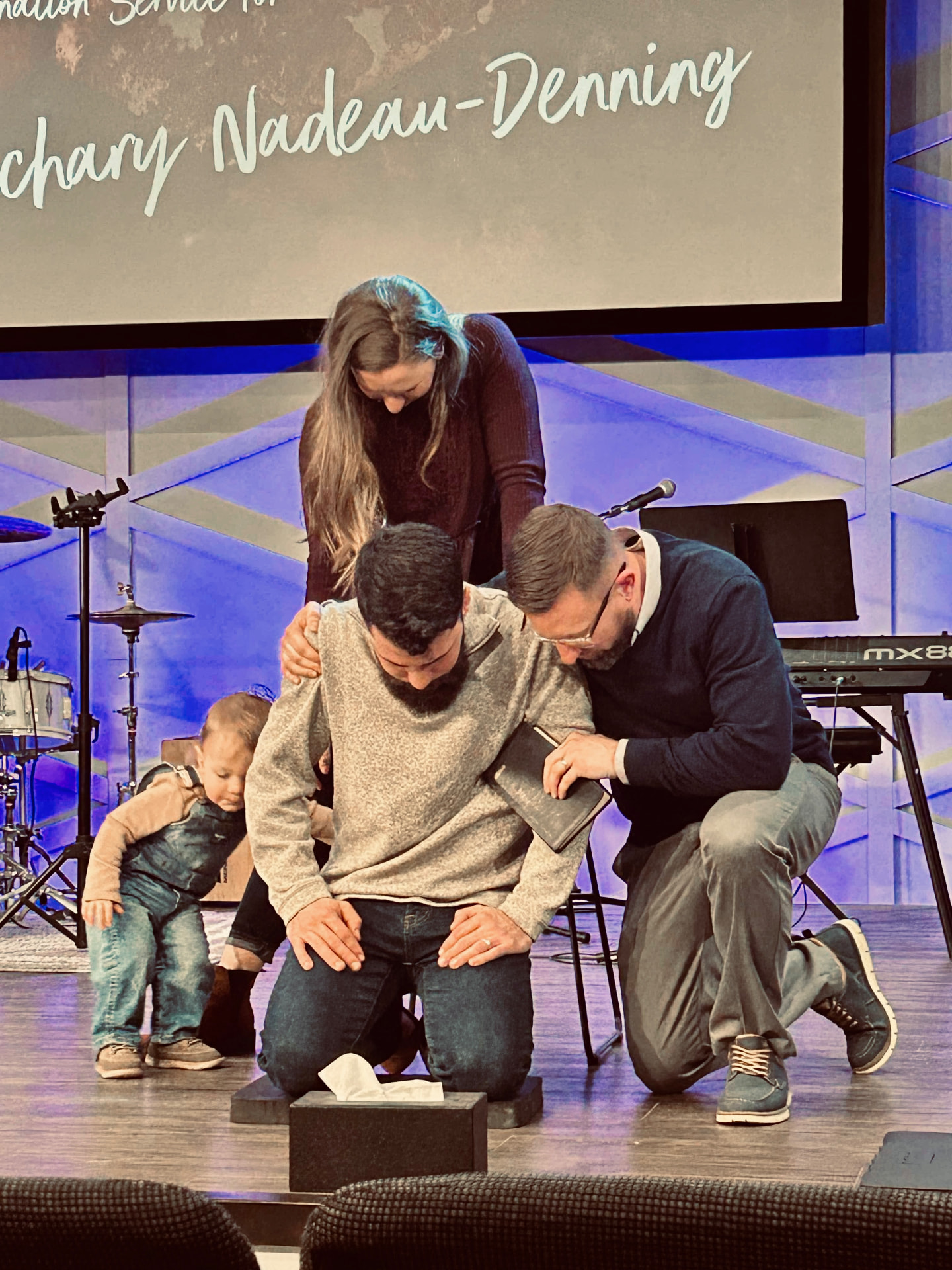 Zachary Denning kneeling in prayer during his ordination service, with two pastors laying hands on him as his name shows on the screen above